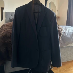 Mörkblå kavaj från Angelico i ull - Snygg mörkblå kavaj från Angelico, tillverkad i Italien av 100% ull. Klassisk modell med två knappar framtill, tre fickor och slits baktill. Perfekt för dig som vill ha en stilren och tidlös look.