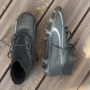 Svarta Nike fotbollsskor med snörning - Säljer ett par svarta Nike fotbollsskor med vit swoosh på sidan. Skorna har snörning, högre strumpliknande skaft och dobbar för bra grepp på planen. Snygg och sportig design som passar perfekt för matcher och träning.
