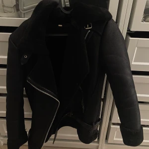 Svart jacka - Snygg svart jacka med bred krage, detaljer i skinn och päls, samt bälte i midjan. Jackan har dragkedja framtill och uppvikta ärmslut. Perfekt för dig som vill ha en cool och varm look.