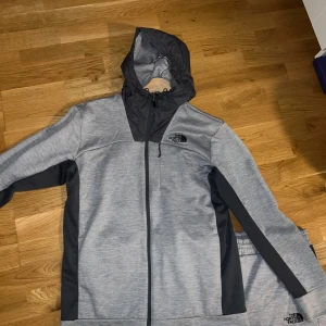 Grå träningsset från The North Face - Säljer ett grått träningsset från The North Face med matchande hoodie och byxor. Hoodien har svart huva och svarta detaljer längs sidorna och ärmarna, samt dragkedja framtill. Byxorna har resår i midjan och logga på låret. Perfekt för gymmet eller chill. Knappt använt