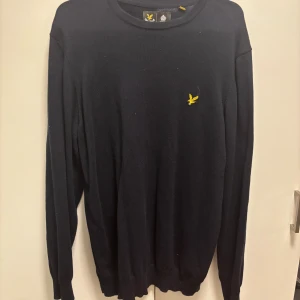 Mörkblå stickad tröja Lyle & Scott - Snygg mörkblå stickad tröja från Lyle & Scott med klassisk rund halsringning och gul broderad logga på bröstet. Perfekt för lager-på-lager och passar till både jeans och chinos.