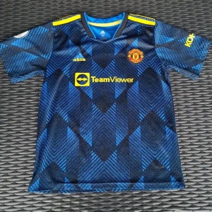 Manchester United Varane #19 bortatröja - Säljer en officiell Manchester United bortatröja från Adidas med Varane och nummer 19 på ryggen. Tröjan är blå med mörkblå geometriskt mönster, gula detaljer och klubbmärke på bröstet. Premier League-logga på ärmen. Tillverkad i lätt polyester, kortärmad och med rund hals.