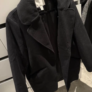 Svart kappa med klassisk krage - Stilren svart kappa från H&M med klassisk krage och raka fickor. Jackan har en tidlös design och är perfekt för dig som gillar en clean och enkel look. Långärmad och fodrad för extra komfort. Säljer för 600kr men kan låta den gå för 500 via snabbaffär.