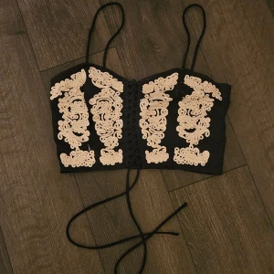 Svart korsettopp med broderi och snörning - Säljer en svart korsettopp med tunna axelband och snygg snörning framtill. Toppen är från min mamma och är köpt runt 2000 som är knappt använd. Toppen har detaljerade beige broderier på framsidan som ger en unik look. Perfekt att styla med jeans eller kjol för en trendig outfit. 