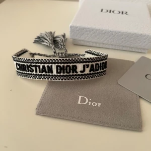 Christian Dior J'Adior armband svart/vit - Snyggt armband från Christian Dior med texten 'J'Adior' broderad i svart på vit bakgrund. Armbandet har justerbar knytning med tofsar och levereras med originalask, dustbag och äkthetskort. Perfekt accessoar för att lyfta din outfit.