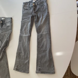 Grå bootcut jeans från Gina Tricot - Snygga grå jeans från Gina Tricot i bootcut-modell med dekorativa bakfickor och silverfärgade knappar. Jeansen har klassisk femficksdesign, normal passform och är tillverkade i mjukt denimtyg. Perfekt för dig som gillar en cool och avslappnad stil. Det finns inga defekter, men dem har används.