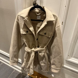 Beige jacka från Zara med bälte - Stilren beige jacka från Zara med bälte i midjan, stora bröstfickor med knappar och klassisk krage. Jackan har långa ärmar och är tillverkad i ett mjukt ullblandat material. Perfekt för lager-på-lager och en chill look.