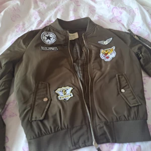 Olivgrön bomberjacka med patches - Snygg olivgrön bomberjacka från S. Oliver med coola broderade patches, bland annat tiger, stjärna och US Army. Jackan har dragkedja, ribbade muddar och två fickor med tryckknappar. Perfekt för dig som vill sticka ut med en streetig look.