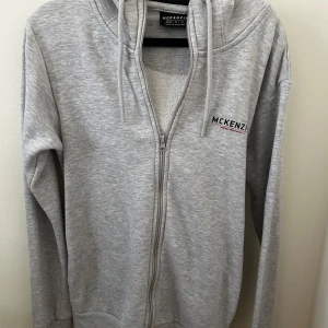 Grå hoodie från McKenzie med dragkedja - Säljer en ljusgrå hoodie från McKenzie i storlek XS. Tröjan har huva med snörning, dragkedja framtill och diskret logga på bröstet. Tillverkad i mjuk bomullsmix och har långa ärmar. Perfekt för chill dagar eller när du vill vara bekväm.