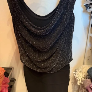 Svart glittrig klänning - Supersnygg svart kortklänning med glittrigt tyg upptill och draperad urringning. Klänningen har en figurnära passform och är perfekt för dig som vill sticka ut. Materialet känns mjukt och stretchigt, och den glittriga ytan ger en extra festlig touch.