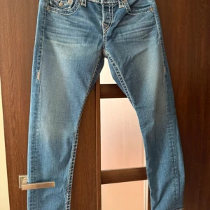 True Religion ljusblå raka jeans - Säljer ett par ljusblå jeans från True Religion med klassiska kontrastsömmar och stora bakfickor med lock och knapp. Jeansen har rak i modell och är tillverkade i mjukt denim med snygg slitning framtill. Perfekta för dig som gillar en avslappnad och trendig look.
