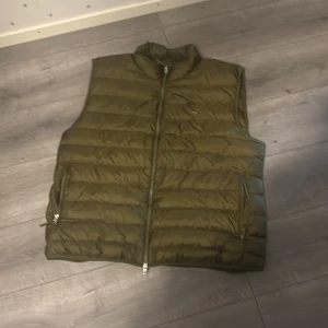 Olivgrön dunväst från GANT XXXL - Snygg olivgrön dunväst från GANT i storlek XXXL. Västen har quiltad design, hög krage och dragkedja framtill samt två fickor med dragkedja. Perfekt för lager-på-lager och chill höst- eller vårstil.
