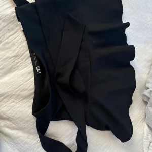 Svart omlottklänning från Zara - Säljer en svart omlottklänning från Zara i storlek S. Klänningen har knytband i midjan och en stilren, enkel design. Perfekt för dig som gillar minimalistisk stil och vill ha något som känns både trendigt och klassiskt. kjolen. Har däremot också inbyggda shorts.