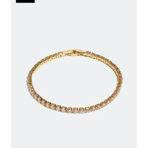 Guldigt tennisarmband med stenar - Ett elegant tennisarmband i guldton med gnistrande klara stenar längs hela bandet. Armbandet har en klassisk, tunn design och smidig låsmekanism. Perfekt för dig som gillar stilrena och glittriga accessoarer som sticker ut på handleden.