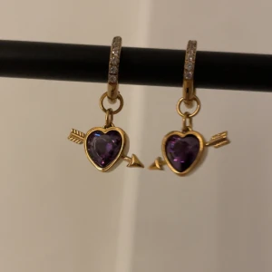 Guldörhängen med lila hjärtan och pil - Superfina örhängen i guldton med gnistrande stenar på ringen och hängande hjärtformad lila sten. Hjärtat är dekorerat med en pil som går igenom, vilket ger en unik och lekfull vibe. Perfekt för dig som gillar detaljer och färg!