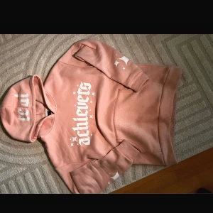 Rosa hoodie med tryck från Real Achievers - Säljer en snygg rosa hoodie från Real Achievers med vit text och stjärnor på bröstet, ärmen och huvan. Tröjan har känguruficka och mjukt material, perfekt för chill dagar. Oversized fit för extra komfort.