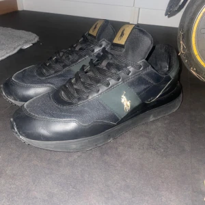Svarta sneakers från Polo Ralph Lauren - Snygga svarta sneakers från Polo Ralph Lauren med guldfärgade detaljer och logga på sidan och hälen. Skorna har rund tå, platt sula och snörning. Ovandelen är i mesh och skinn för en cool och sportig look. Perfekta för dig som gillar stilrena och klassiska sneakers. MYCKET BRA SKICK