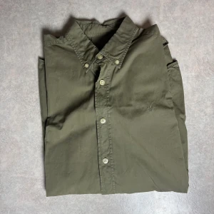 Olivgrön skjorta från Morris M - Snygg olivgrön skjorta från Morris med klassisk krage och knäppning framtill. Skjortan har en broderad lilja på bröstet och är tillverkad i mjuk bomull. Perfekt för dig som gillar stilrena och tidlösa plagg.