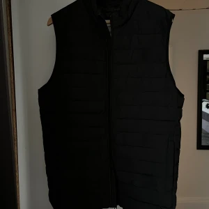 Svart dunväst från Jack & Jones - Säljer en svart dunväst från Jack & Jones i storlek S. Västen har quiltad design, hög krage och dragkedja framtill. Perfekt att slänga över en hoodie för extra värme. Enkel och stilren look som funkar till det mesta. Har använts 1-2 gånger och är i helt nytt skick. 