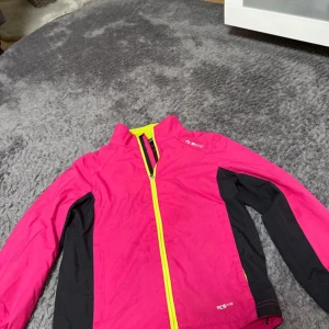 Rosa vindjacka från SOC med gula detaljer - Sportig rosa vindjacka från SOC med svarta paneler längs sidor och ärmar samt gul dragkedja. Jackan har hög krage med meshfoder och långa ärmar. Perfekt för utomhusaktiviteter och träning.