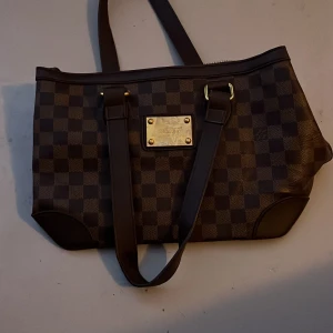 Louis Vuitton rutig axelväska brun - Snygg axelväska från Louis Vuitton i klassiskt rutigt mönster. Väskan är brun med mörkare bruna detaljer och har en guldig metallplatta framtill. Tillverkad i skinn med två långa axelremmar och dragkedja upptill. Perfekt storlek för att få plats med allt du behöver.