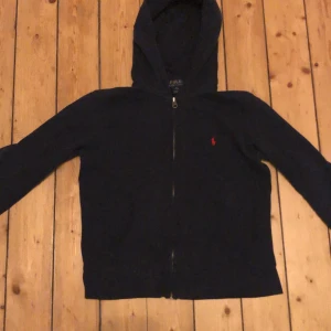 Mörkblå hoodie från Polo Ralph Lauren - Mörkblå hoodie från Polo Ralph Lauren med dragkedja framtill och klassisk huva. Tröjan har lång ärm och en liten röd broderad logga på bröstet. Perfekt för en chill och stilren look.