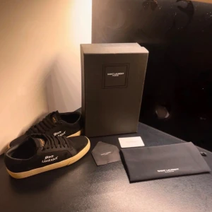 Svarta Saint Laurent sneakers - Snygga svarta sneakers från Saint Laurent med vit logga på sidan och gulaktig platt sula. Skorna har klassisk snörning,  Perfekta för dig som vill ha en stilren och lyxig look. Kommer med originalkartong och dustbag.