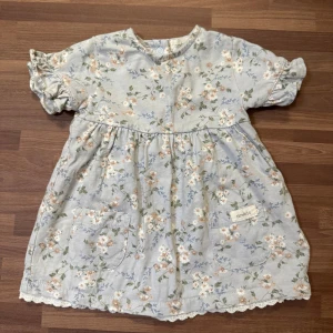 Blommig blå klänning - Supersöt ljusblå klänning från Newbie med blommigt mönster, storlek 68 (4-6mån). Använd fåtal gånger, extremt fint skick helt utan anmärkning. Knappar i ryggen, två fickor framtill och spetskant nedtill.