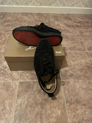 Christian Louboutin svarta sneakers mocka - Säljer ett par svarta sneakers från Christian Louboutin i mocka med klassiska röda sulor och coola nitar på tån. Skorna har snörning och en rund tå, samt en lyxig känsla med sin unika design. Perfekta för dig som vill sticka ut med exklusiva detaljer.