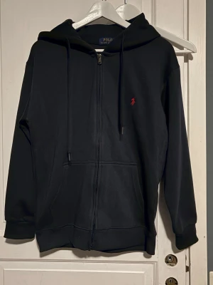 Svart zip hoodie från Polo Ralph Lauren - Klassisk svart hoodie med dragkedja från Polo Ralph Lauren. Har huva med dragsko, fickor framtill och den ikoniska röda logotypen broderad på bröstet. Tillverkad i mjukt sweatshirtmaterial som är skönt att bära. Perfekt för en avslappnad och stilren look. Storlek M men passar för någon som har S.