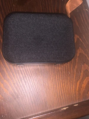 Svart bärbar högtalare med tygfront - Snygg bärbar högtalare i svart med rundade hörn och tygfront. Baksidan är i matt plast och den har en praktisk rem i brunt tyg. Utrustad med AUX-ingång och USB-C-port för laddning. Perfekt för att ta med sig musiken överallt.