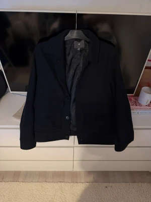 Svart overshirt från H&M - Stilren svart overshirt från H&M med klassisk krage och stora fickor framtill. Jackan har knäppning med knappar och är helsvart, perfekt för lager på lager. Tillverkad i ett mjukt material som känns bekvämt och passar till många olika looks. Storlek S. Ordpris: 399kr säljer för 250kr