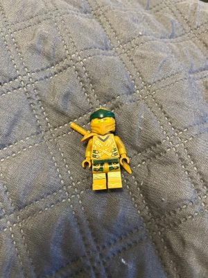 LEGO Ninjago guldig ninjafigur - Säljer en LEGO Ninjago minifigur i guld med gröna detaljer och ett grönt band runt huvudet. Figuren har ett svärd på ryggen och coola tryck på kroppen. Perfekt till samlingen eller för att leka ninjaäventyr!