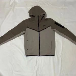 Nike grå hoodie med dragkedja - Snygg grå hoodie från Nike med hel dragkedja framtill och svart detalj på ärmen. Tröjan har huva, två fickor och en svart Nike-logga på bröstet. Tillverkad i mjukt material som passar perfekt till en avslappnad stil.