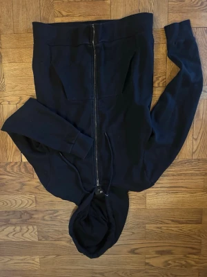 Svart hoodie med dragkedja - Svart hoodie med hel dragkedja framtill och två stora fickor. Klassisk huva med dragsko och ribbade muddar vid ärmslut och nederkant. Perfekt för en avslappnad och stilren look. Materialet känns mjukt och bekvämt, troligen bomullsblandning.