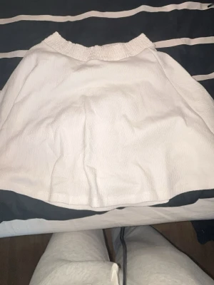 Vit kortkjol med resårmidja - Säljer en enkel och stilren vit kortkjol med resår i midjan. Kjolen har en lätt utställd form och är gjord i ett mjukt material som känns bekvämt att bära. Perfekt att matcha med en crop top eller hoodie.