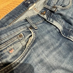Blå jeans från Gant - Säljer ett par klassiska blå jeans från Gant med raka ben och normal passform. Jeansen har fem fickor, bälteshällor och Gant-logga på myntfickan. Materialet är slitstarkt denim i bomull och färgen är mellanblå med lätt tvättad look. Byxorna är omsydda imellan bena eftersom de sprack men inget som syns utifrån! Även längst ner på benen