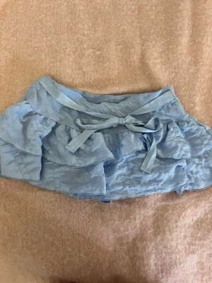 Ljusblå volangkjol med rosett - Supersöt ljusblå kortkjol med volanger och en dekorativ rosett i midjan. Kjolen har en luftig och lätt struktur, perfekt för varma dagar. Passar dig som vill ha en feminin och trendig look. Har inbyggda shorts