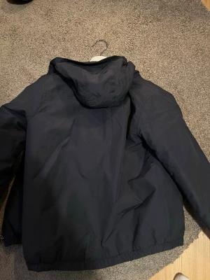 Svart vindjacka från Lyle & Scott - Svart vindjacka från Lyle & Scott med huva och enkel design. Jackan är tillverkad i syntetmaterial och har en klassisk passform. Perfekt för blåsiga dagar och enkel att matcha med olika outfits.