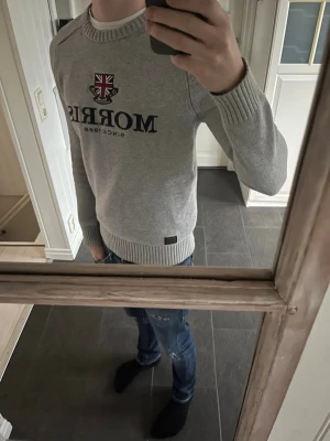 Morris tröja - Snygg grå tröja från Morris med broderad logga och brittisk flagga på bröstet. Tröjan har rund halsringning, ribbade muddar och en klassisk passform. Perfekt för en stilren och avslappnad look. Modell: 176cm