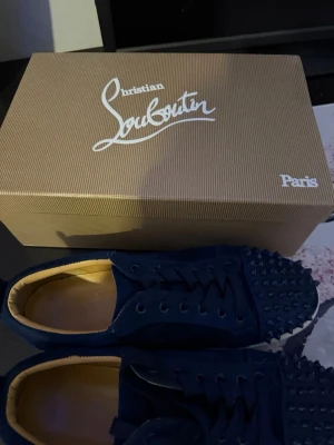 Blå sneakers från Christian Louboutin - Säljer ett par blå sneakers från Christian Louboutin med ikoniska röda sulor och coola nitar på tån. Skorna har snörning och är tillverkade i mocka för en lyxig känsla. Perfekta för dig som vill sticka ut med stilrena detaljer. Skorna är äkta bara att skriva om du har frågor.