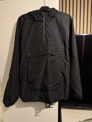 Svart vindjacka med huva och ficka - Snygg svart vindjacka med huva och stor magficka med knappar. Jackan har dragsko i nederkant och vid huvan samt half zip framtill. Perfekt för blåsiga dagar och enkel att matcha till olika outfits.