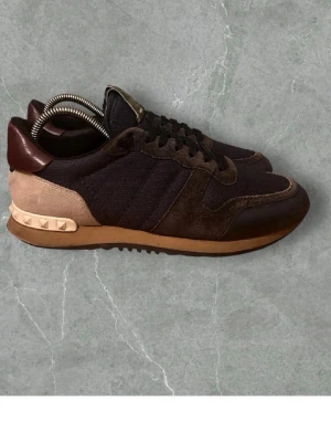 Valentino Rockrunner sneakers brun - Snygga Valentino Rockrunner sneakers i mörkbrunt och beige med detaljer i skinn, mocka och textil. Skorna har klassisk snörning, nitar på hälen och en kontrasterande sula. Perfekta för dig som gillar exklusiva och trendiga sneakers.