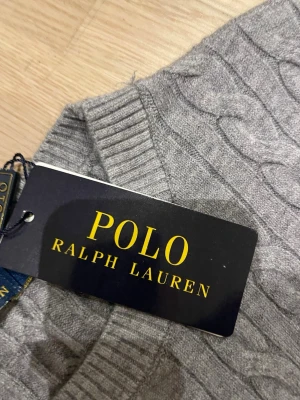 Grå kabelstickad tröja Polo Ralph Lauren - Snygg grå kabelstickad tröja från Polo Ralph Lauren med rund halsringning och diskret broderad logga på bröstet. Tröjan har ribbade muddar vid ärmslut och nederkant. Perfekt för lager på lager och en clean look.