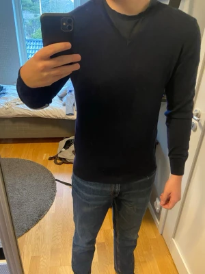 Mörkblå stickad tröja herr - Snygg mörkblå stickad tröja med rund halsringning och långa ärmar. Tröjan har ribbade muddar vid ärmslut och nederkant för en tajtare passform. Perfekt att bära över en t-shirt eller skjorta för en clean och stilren look. 100% merinoull. Väldigt fint skick!