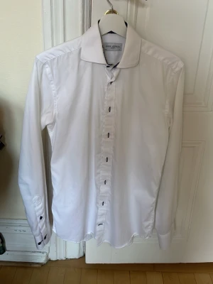 Vit skjorta från John Henric - Klassisk vit skjorta från John Henric med lång ärm och svarta knappar. Skjortan har en stilren design och är tillverkad i 100% bomull, perfekt för en clean och snygg look. Passar till många olika outfits och tillfällen.