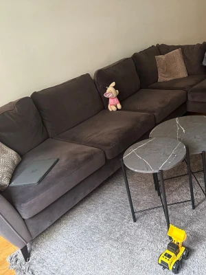 Mörkgrå hörnsoffa i manchester tyg - Stor och mysig mörkgrå hörnsoffa i tyg med flera lösa ryggkuddar. Soffan har en modern design och passar perfekt för större vardagsrum. Inkluderar matchande kuddar i beige och grått. Perfekt för filmkvällar eller häng med vänner.