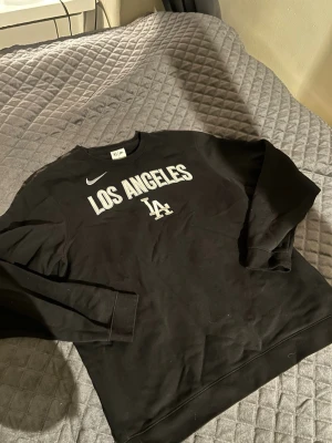 Svart Nike Los Angeles sweatshirt - Säljer en svart sweatshirt från Nike med broderad text 'Los Angeles' och LA-logga på bröstet. Tröjan har rund halsringning och långa ärmar. Perfekt för dig som gillar sportig streetstyle och vill ha något bekvämt och snyggt.