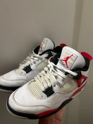 Nike Air Jordan 4 Retro Fire Red - Nike Air Jordan 4 Retro Fire Red med vit ovandel i skinn, svarta och röda detaljer samt meshpaneler på sidorna. Klassisk Jumpman-logga på plösen och hälen. Skorna har snörning och en chunky sula med svart och röd färg. Perfekt för dig som vill sticka ut med ikoniska sneakers.