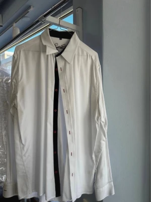 Vit skjorta Arnold Palmer XL - Säljer en vit skjorta från Arnold Palmer i storlek XL. Skjortan har svarta detaljer längs knappslån och röda knappar som ger en snygg kontrast. Klassisk krage och lång ärm, perfekt till jeans eller chinos.